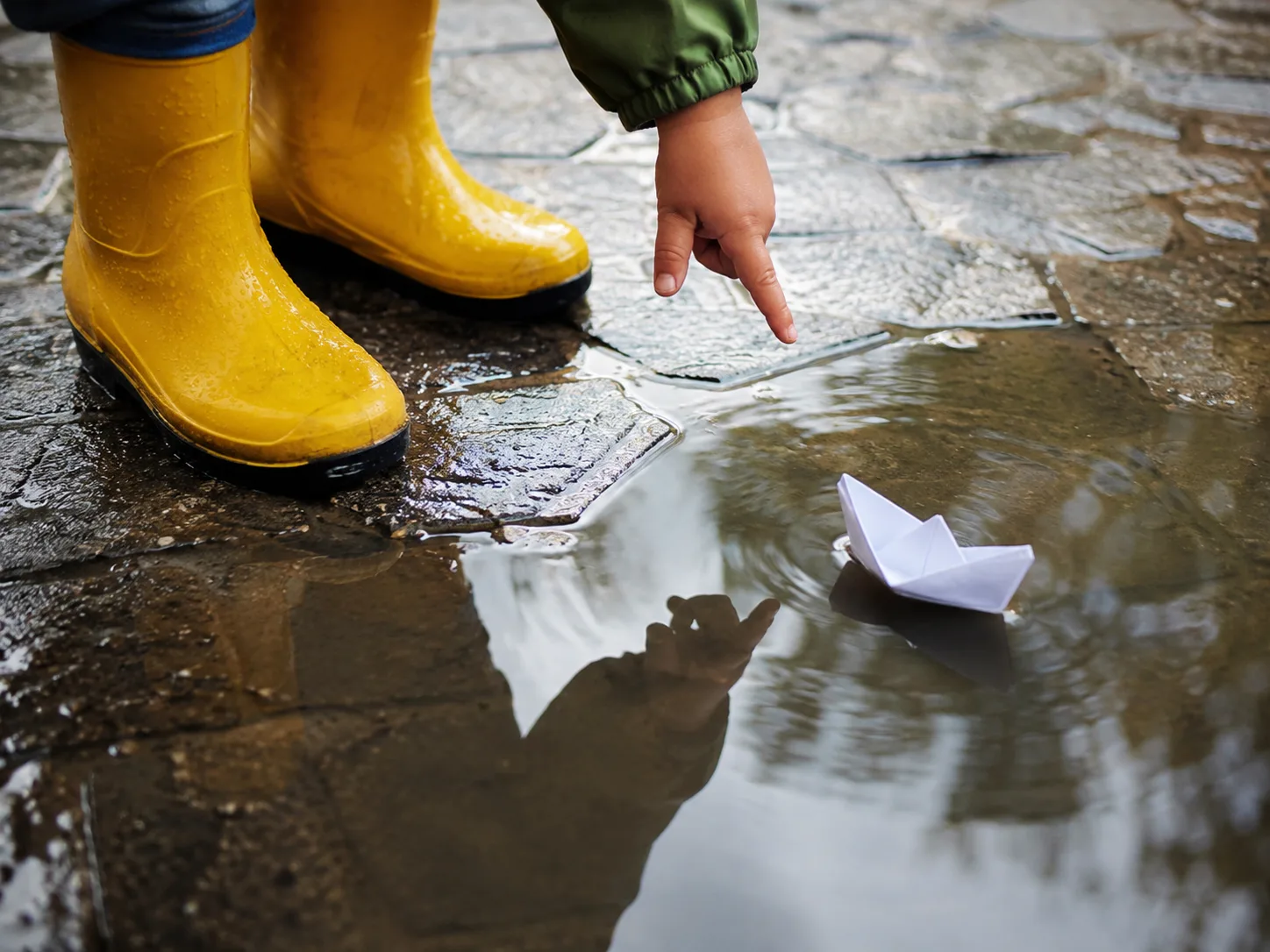 Child in yellow rain boots pointing at a paper boat floating in a rain puddle on wet stone pavement.