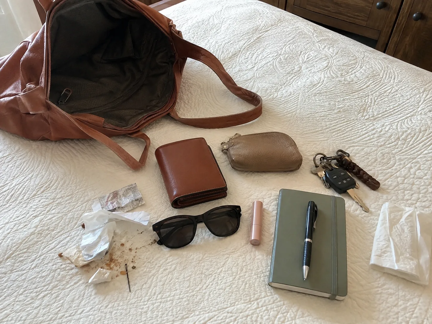 Open everyday bag emptied onto a bed with a wallet, keys, sunglasses, notebook, pen, small pouch, receipts and rubbish ready to sort.