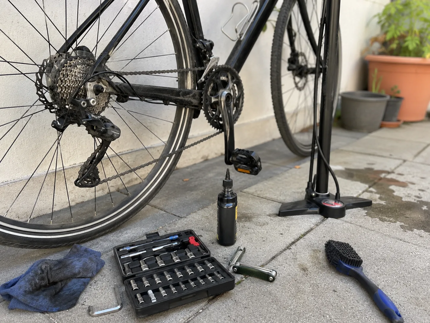 Black bicycle being repaired outdoors with a pump, lubricant, cleaning brush, cloth, and bike tools arranged beside the rear wheel.